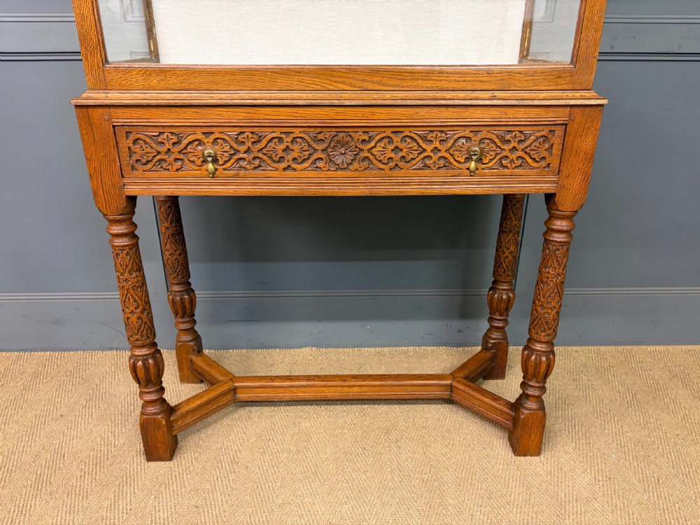 A tall carved oak display cabinet featuring three glass shelves, glazed sides, a carved frieze drawer, and carved turned legs. View IX