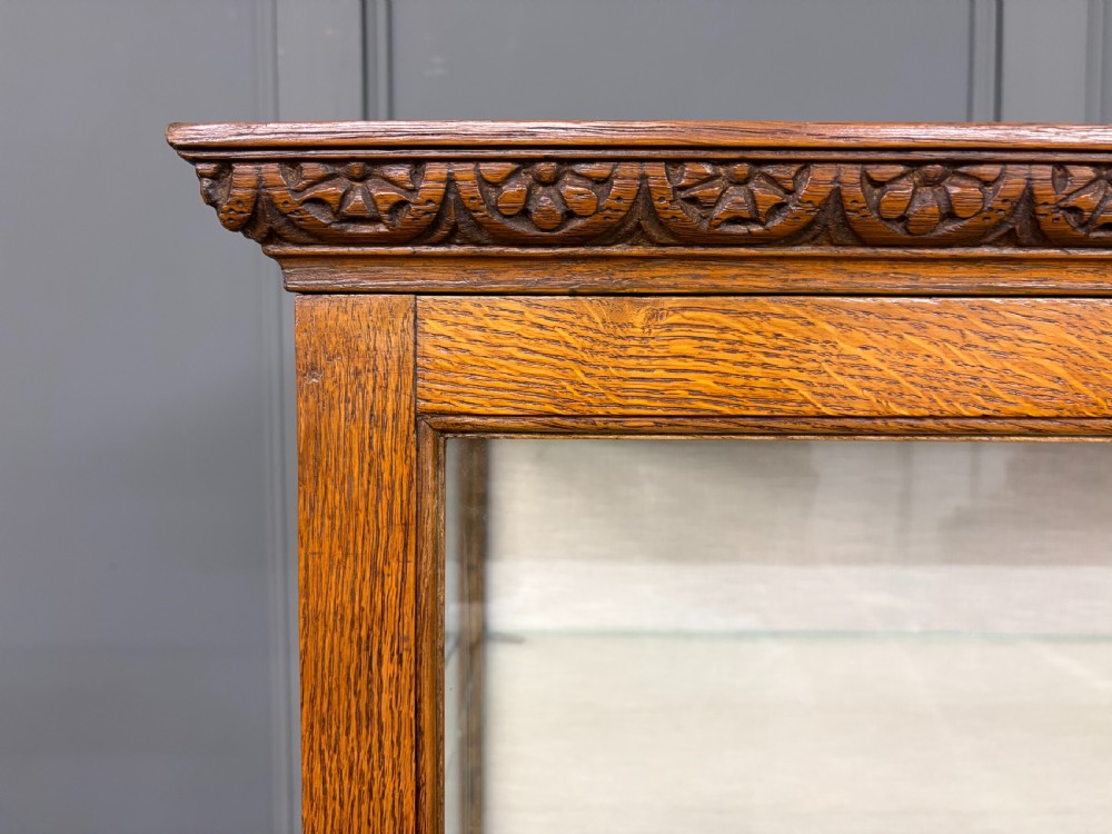 A tall carved oak display cabinet featuring three glass shelves, glazed sides, a carved frieze drawer, and carved turned legs. View X