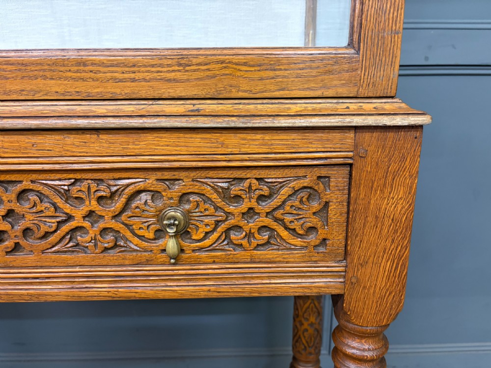 A tall carved oak display cabinet featuring three glass shelves, glazed sides, a carved frieze drawer, and carved turned legs. View XI