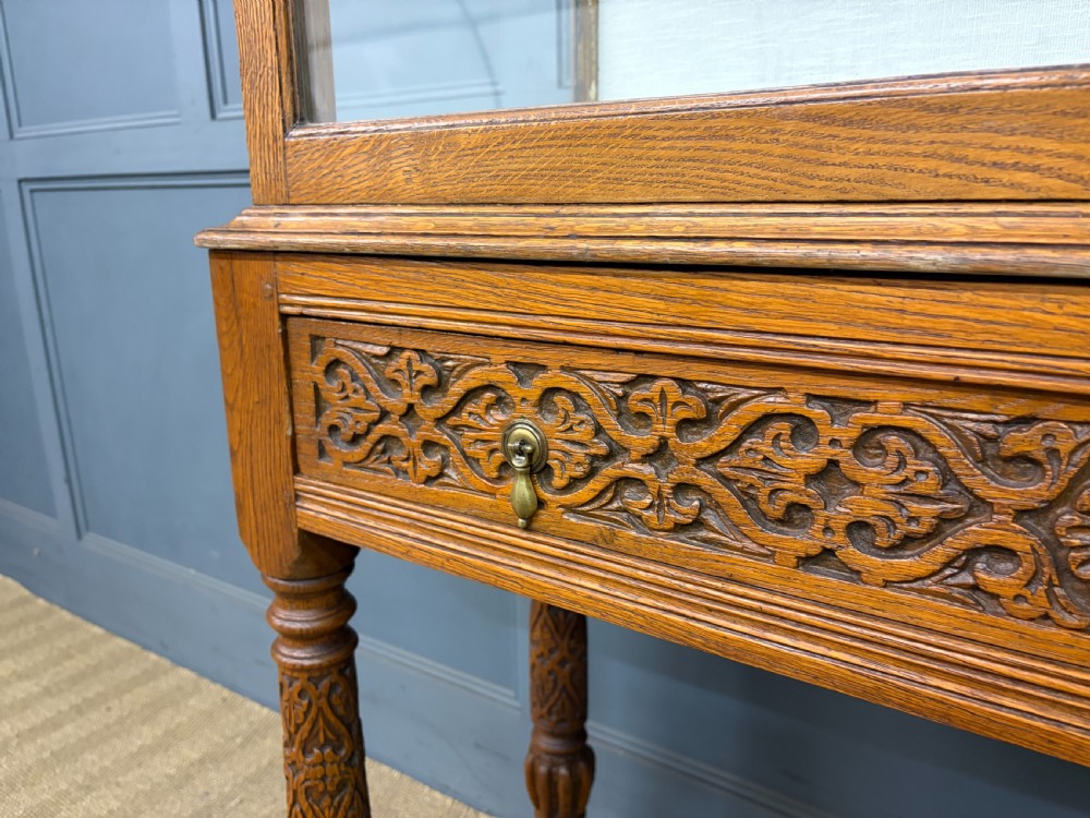 A tall carved oak display cabinet featuring three glass shelves, glazed sides, a carved frieze drawer, and carved turned legs. View XII