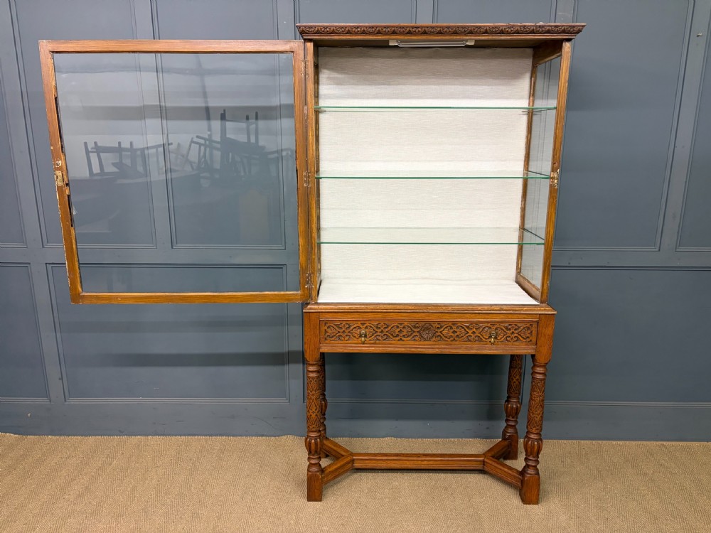 A tall carved oak display cabinet featuring three glass shelves, glazed sides, a carved frieze drawer, and carved turned legs. View XV