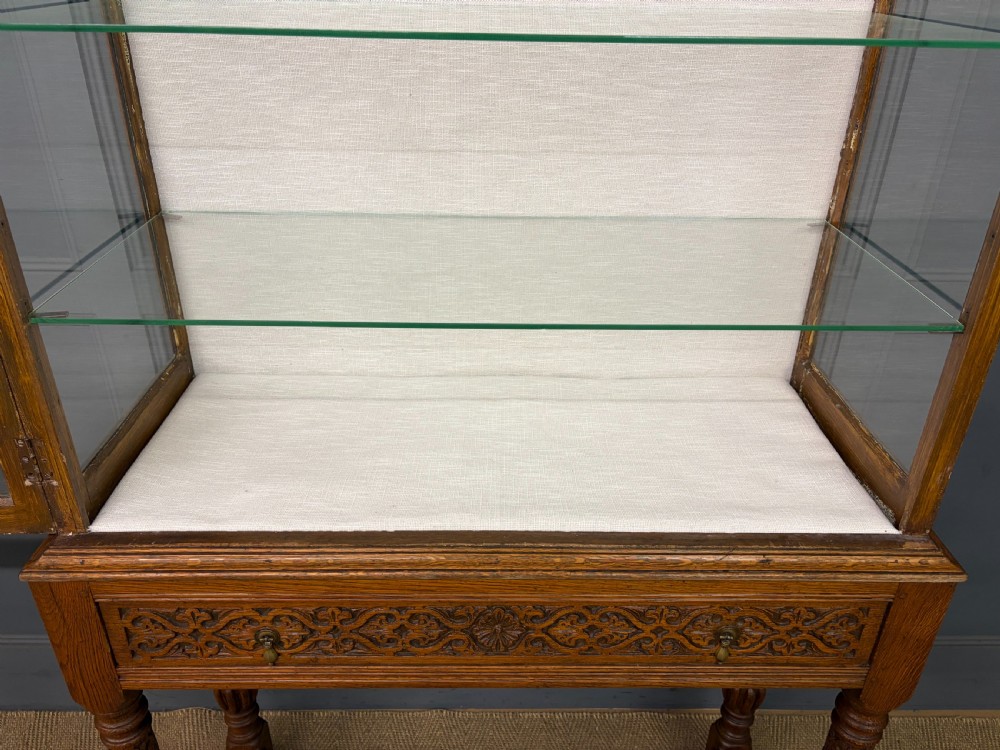 A tall carved oak display cabinet featuring three glass shelves, glazed sides, a carved frieze drawer, and carved turned legs. View XVI