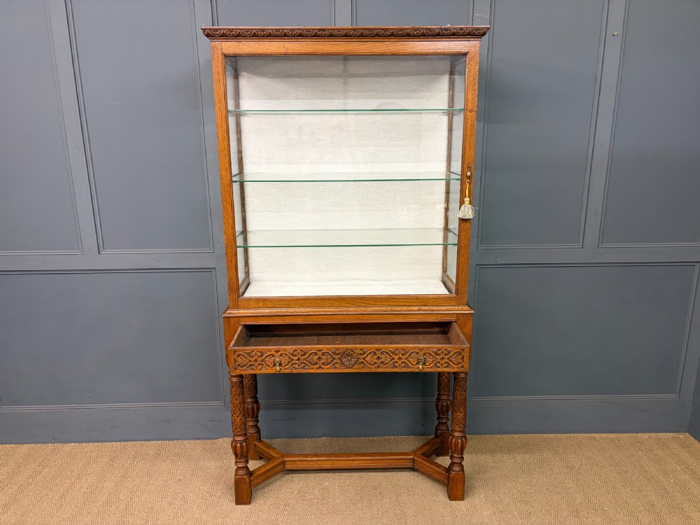 A tall carved oak display cabinet featuring three glass shelves, glazed sides, a carved frieze drawer, and carved turned legs. View XVII