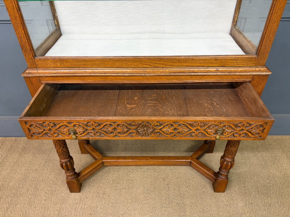 A tall carved oak display cabinet featuring three glass shelves, glazed sides, a carved frieze drawer, and carved turned legs. View XVIII