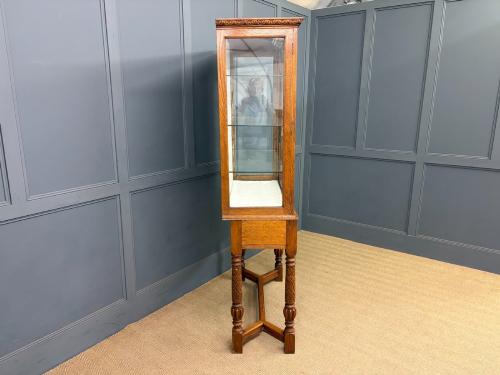A tall carved oak display cabinet featuring three glass shelves, glazed sides, a carved frieze drawer, and carved turned legs. View XX