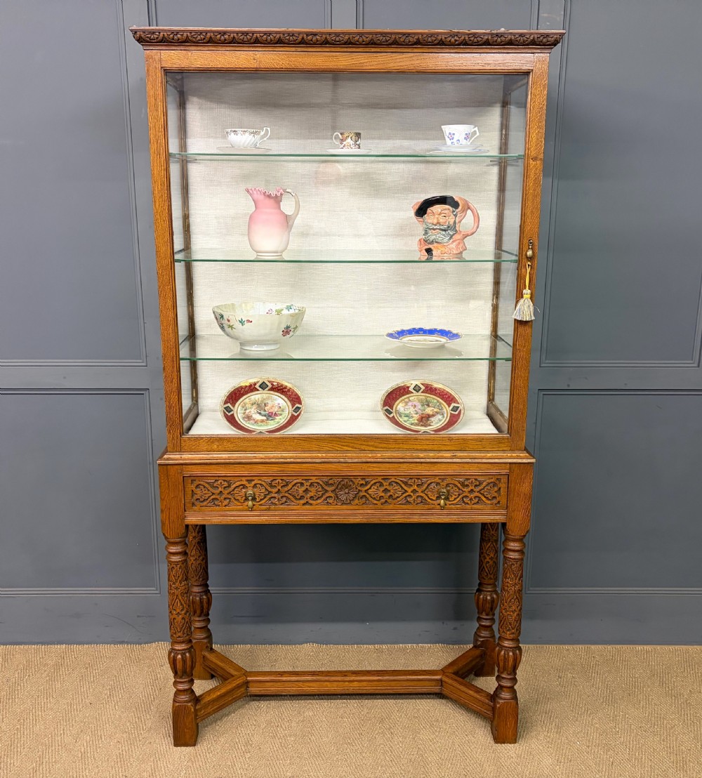 A tall carved oak display cabinet featuring three glass shelves, glazed sides, a carved frieze drawer, and carved turned legs. View XXII