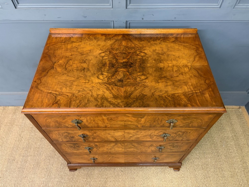 An Art Deco burr walnut chest of four full-width drawers featuring figured veneer panels, decorative brass drop handles, and low bracket feet. View IV