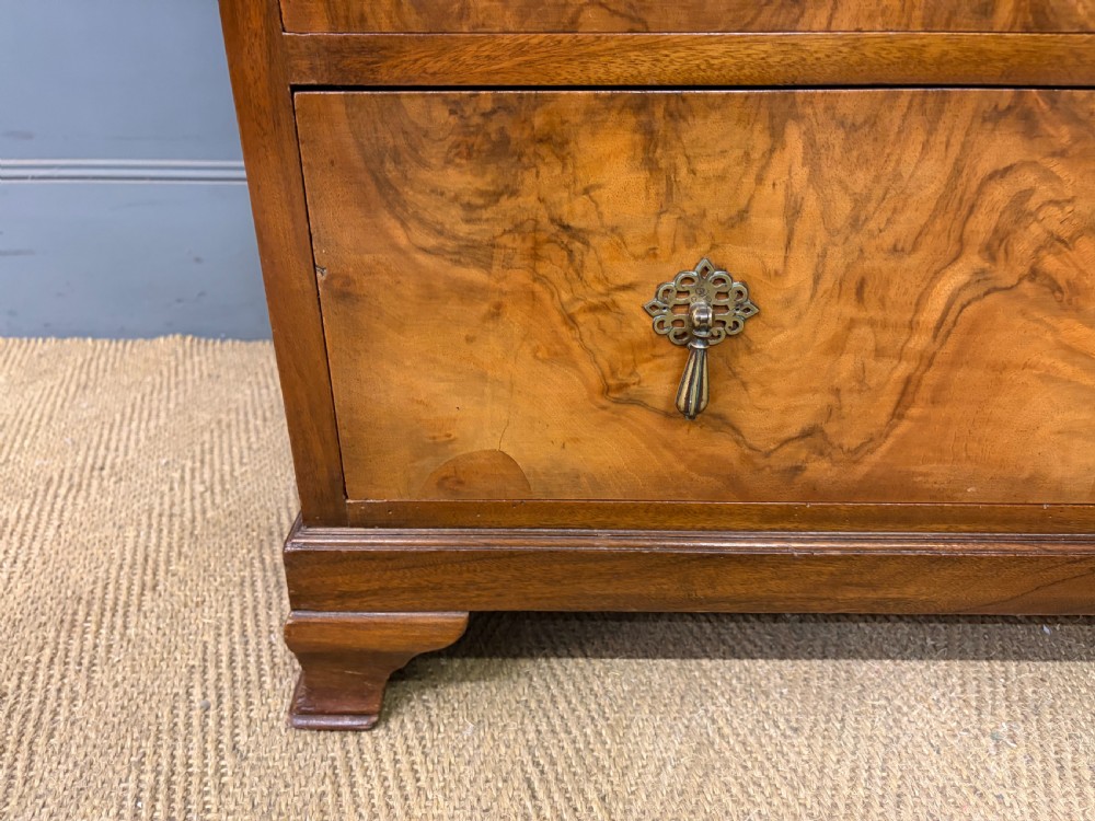 An Art Deco burr walnut chest of four full-width drawers featuring figured veneer panels, decorative brass drop handles, and low bracket feet. View VII