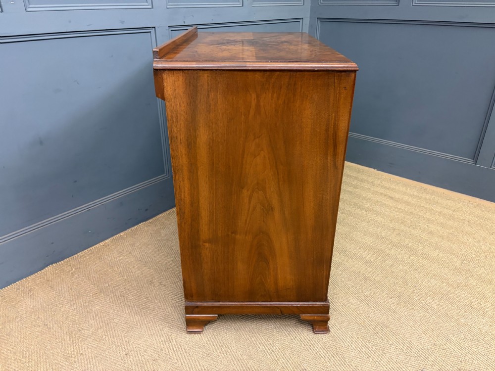 An Art Deco burr walnut chest of four full-width drawers featuring figured veneer panels, decorative brass drop handles, and low bracket feet. View X
