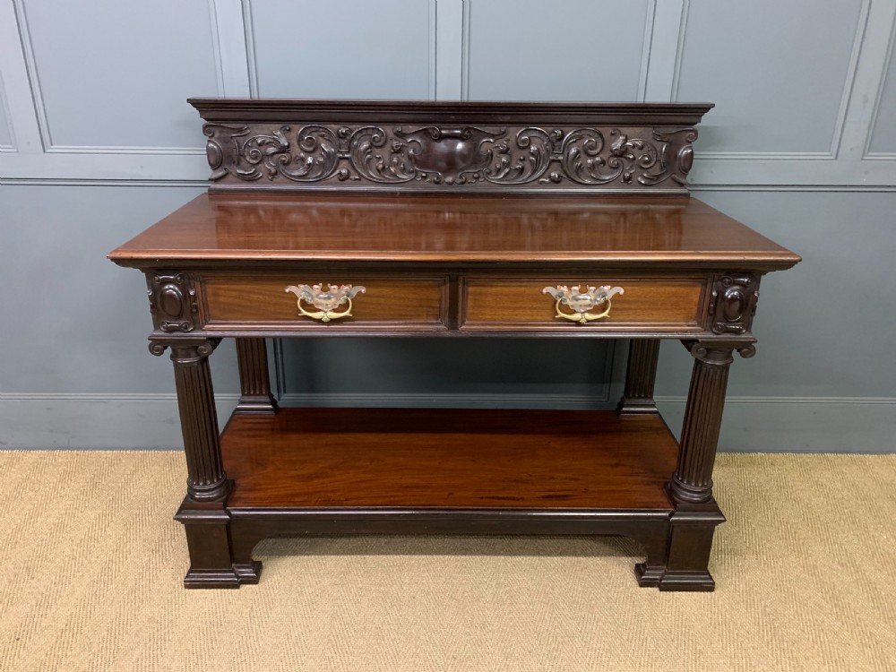 A Victorian dark mahogany buffet featuring a scroll-carved backboard, two drawers with brass handles, fluted front columns, and an open lower tier.