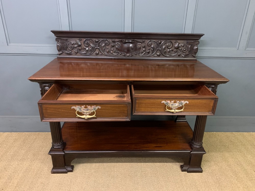 A Victorian dark mahogany buffet featuring a scroll-carved backboard, two drawers with brass handles, fluted front columns, and an open lower tier. View XII