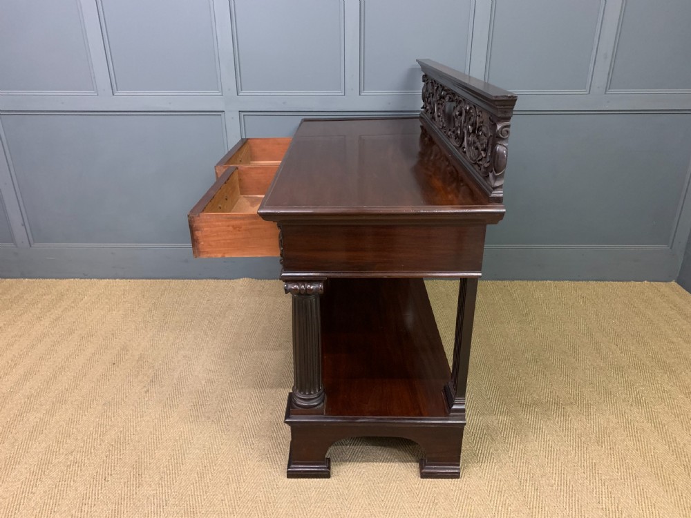 A Victorian dark mahogany buffet featuring a scroll-carved backboard, two drawers with brass handles, fluted front columns, and an open lower tier. View XVII