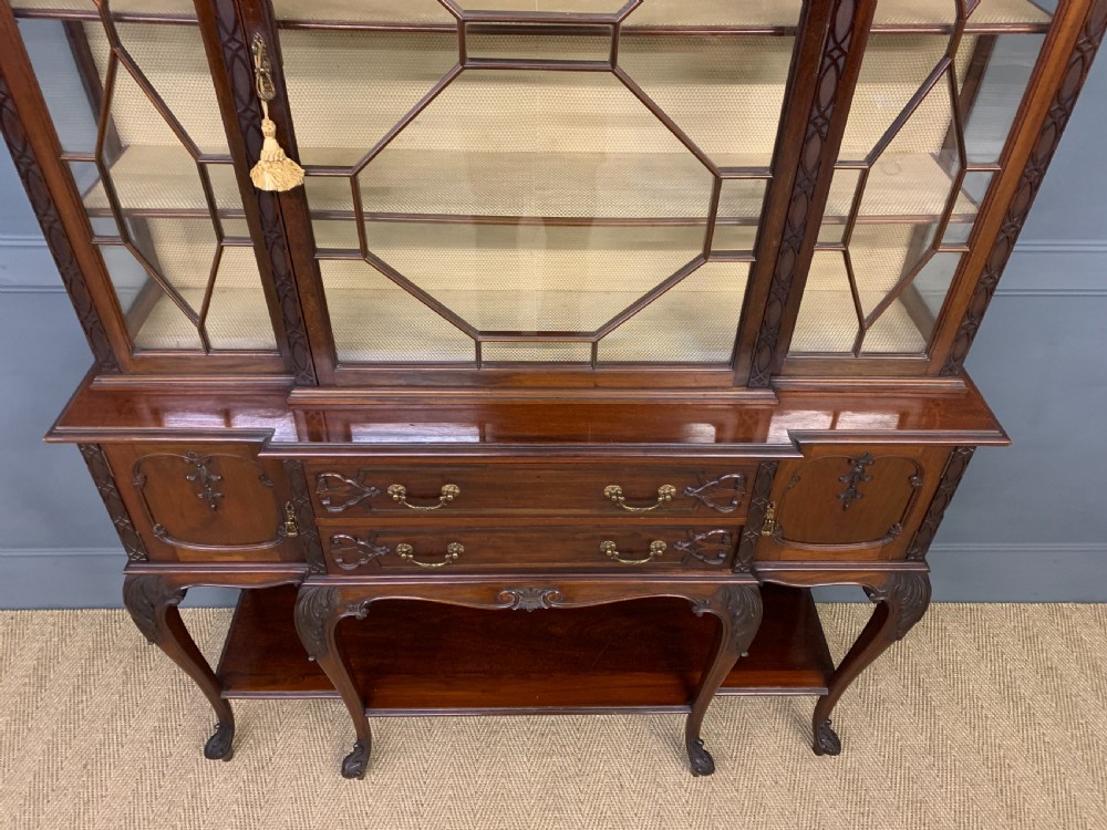 A carved dark mahogany display cabinet featuring astragal glazed doors, lower drawers, carved cabriole legs, and an undertier shelf. View IX