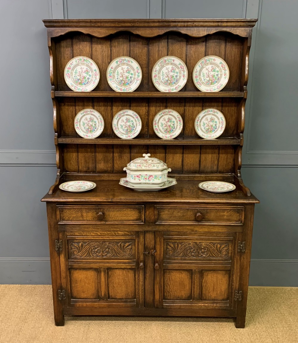 A dark oak Welsh dresser featuring an upper open plate rack with three shelves above a lower cabinet with two drawers and two carved panel doors.
