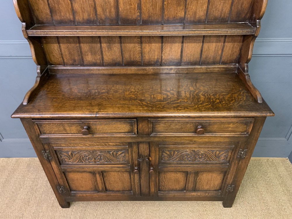 A dark oak Welsh dresser featuring an upper open plate rack with three shelves above a lower cabinet with two drawers and two carved panel doors. View V