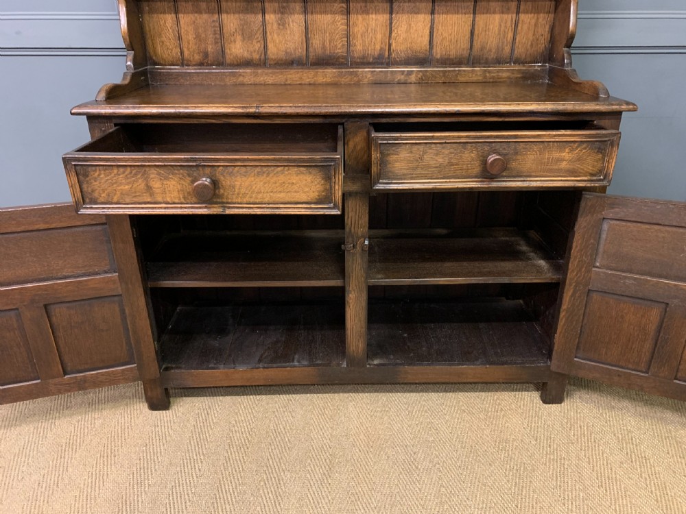 A dark oak Welsh dresser featuring an upper open plate rack with three shelves above a lower cabinet with two drawers and two carved panel doors. View X