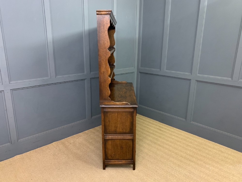 A dark oak Welsh dresser featuring an upper open plate rack with three shelves above a lower cabinet with two drawers and two carved panel doors. View XII