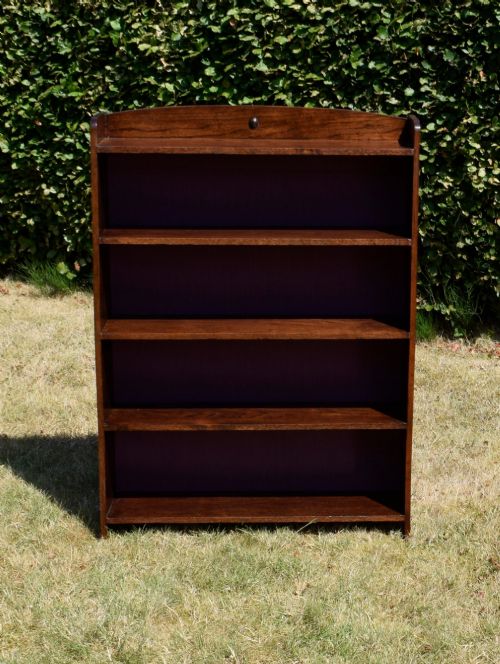 fig 2. An Arts and Crafts solid oak open bookcase featuring five shelves, solid plank sides, an arched gallery top with a central decorative motif, and a purple fabric-lined back panel.