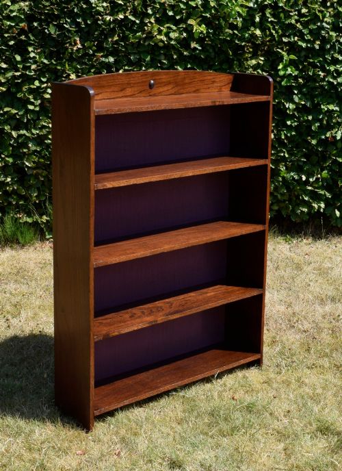 fig 3. An Arts and Crafts solid oak open bookcase featuring five shelves, solid plank sides, an arched gallery top with a central decorative motif, and a purple fabric-lined back panel.