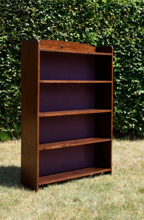 fig 4. An Arts and Crafts solid oak open bookcase featuring five shelves, solid plank sides, an arched gallery top with a central decorative motif, and a purple fabric-lined back panel.