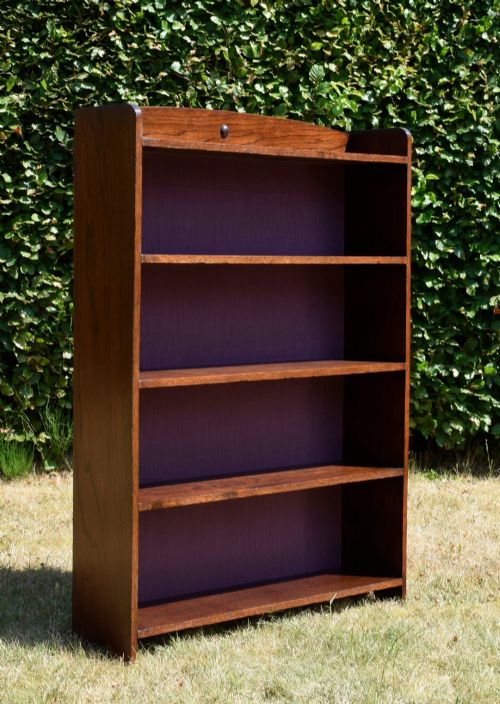 fig 5. An Arts and Crafts solid oak open bookcase featuring five shelves, solid plank sides, an arched gallery top with a central decorative motif, and a purple fabric-lined back panel.