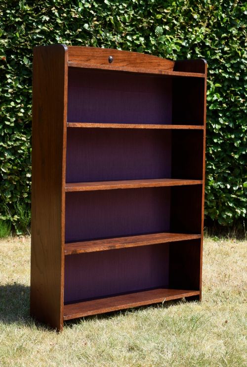 fig 6. An Arts and Crafts solid oak open bookcase featuring five shelves, solid plank sides, an arched gallery top with a central decorative motif, and a purple fabric-lined back panel.