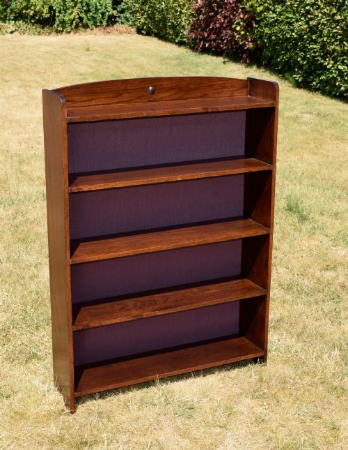 fig 7. An Arts and Crafts solid oak open bookcase featuring five shelves, solid plank sides, an arched gallery top with a central decorative motif, and a purple fabric-lined back panel.