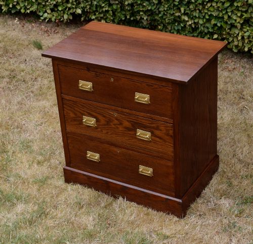 fig 7. A medium-toned oak chest of drawers featuring three drawers with rectangular brass handles resting on a plinth base.