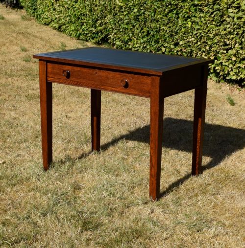 fig 1. A warm brown oak Arts and Crafts writing desk with a dark inset leather top, a single drawer with turned wooden knobs, and four square straight legs.