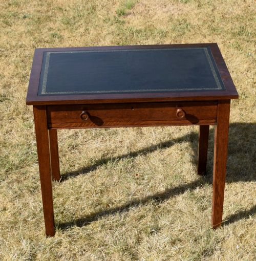 fig 6. A warm brown oak Arts and Crafts writing desk with a dark inset leather top, a single drawer with turned wooden knobs, and four square straight legs.