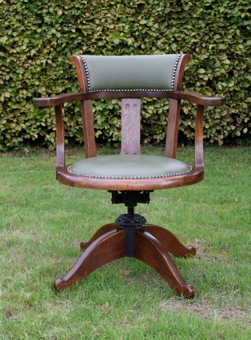 fig 2. An early 20th-century oak swivel desk chair featuring a green leather padded seat and backrest trimmed with metal studs, supported by a four-spoke wooden base with a central iron mechanism.