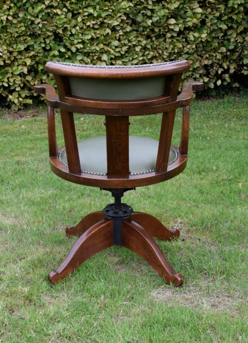 fig 4. An early 20th-century oak swivel desk chair featuring a green leather padded seat and backrest trimmed with metal studs, supported by a four-spoke wooden base with a central iron mechanism.