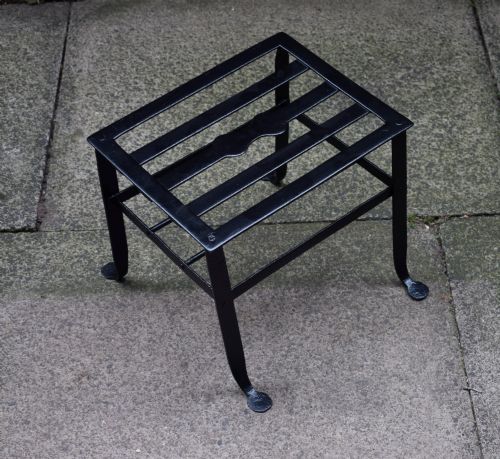 fig 1. A black wrought iron fireplace footman featuring a slatted top with a central decorative cutout handle, resting on four curved legs with flat circular feet.
