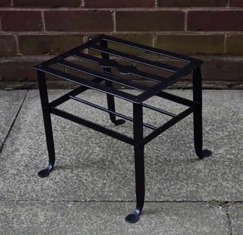 fig 3. A black wrought iron fireplace footman featuring a slatted top with a central decorative cutout handle, resting on four curved legs with flat circular feet.