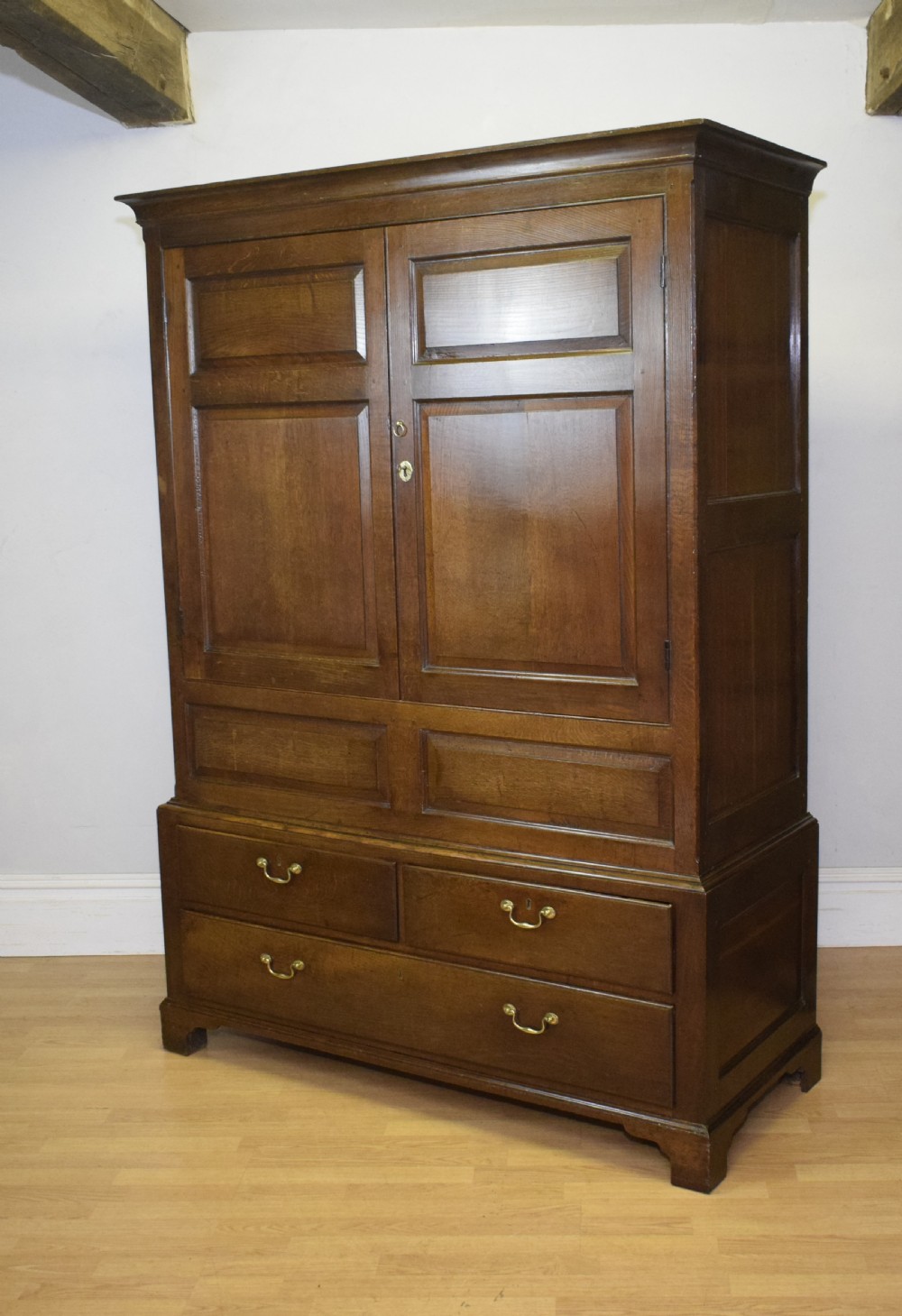 A two-piece Georgian oak press cupboard featuring twin fielded panel doors over a three-drawer base with brass handles.