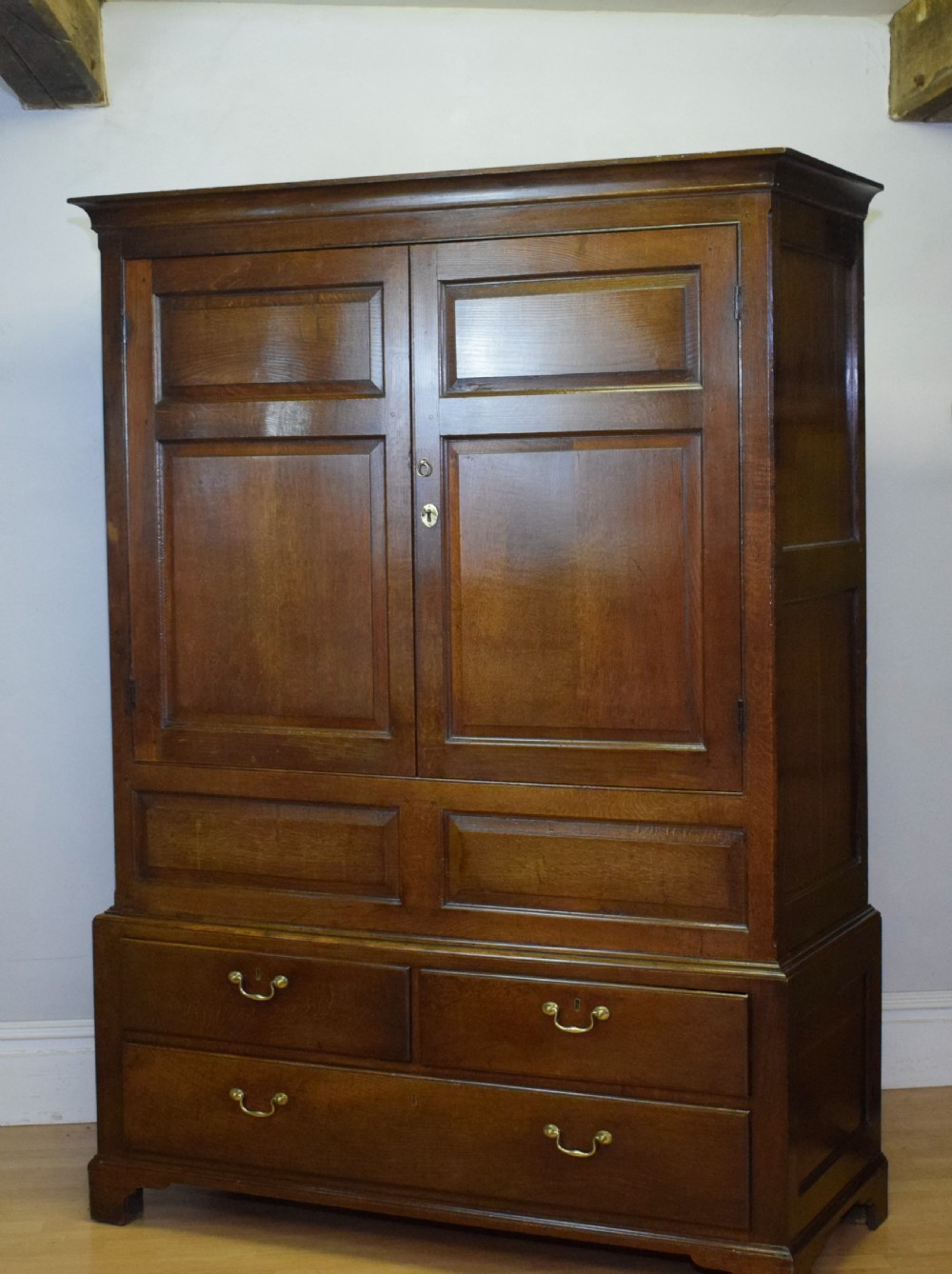 A two-piece Georgian oak press cupboard featuring twin fielded panel doors over a three-drawer base with brass handles. View VII
