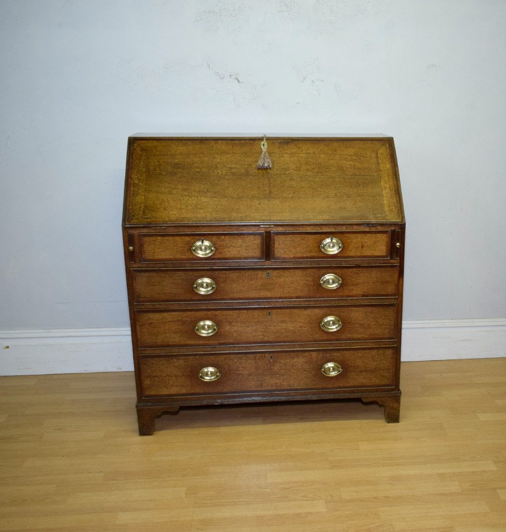 A Georgian oak bureau featuring a slanted fall-front lid above four graduated drawers fitted with brass oval handles and raised on bracket feet. View V