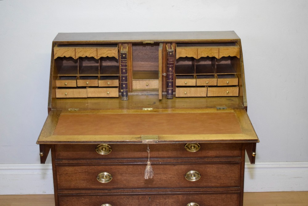 A Georgian oak bureau featuring a slanted fall-front lid above four graduated drawers fitted with brass oval handles and raised on bracket feet. View IX