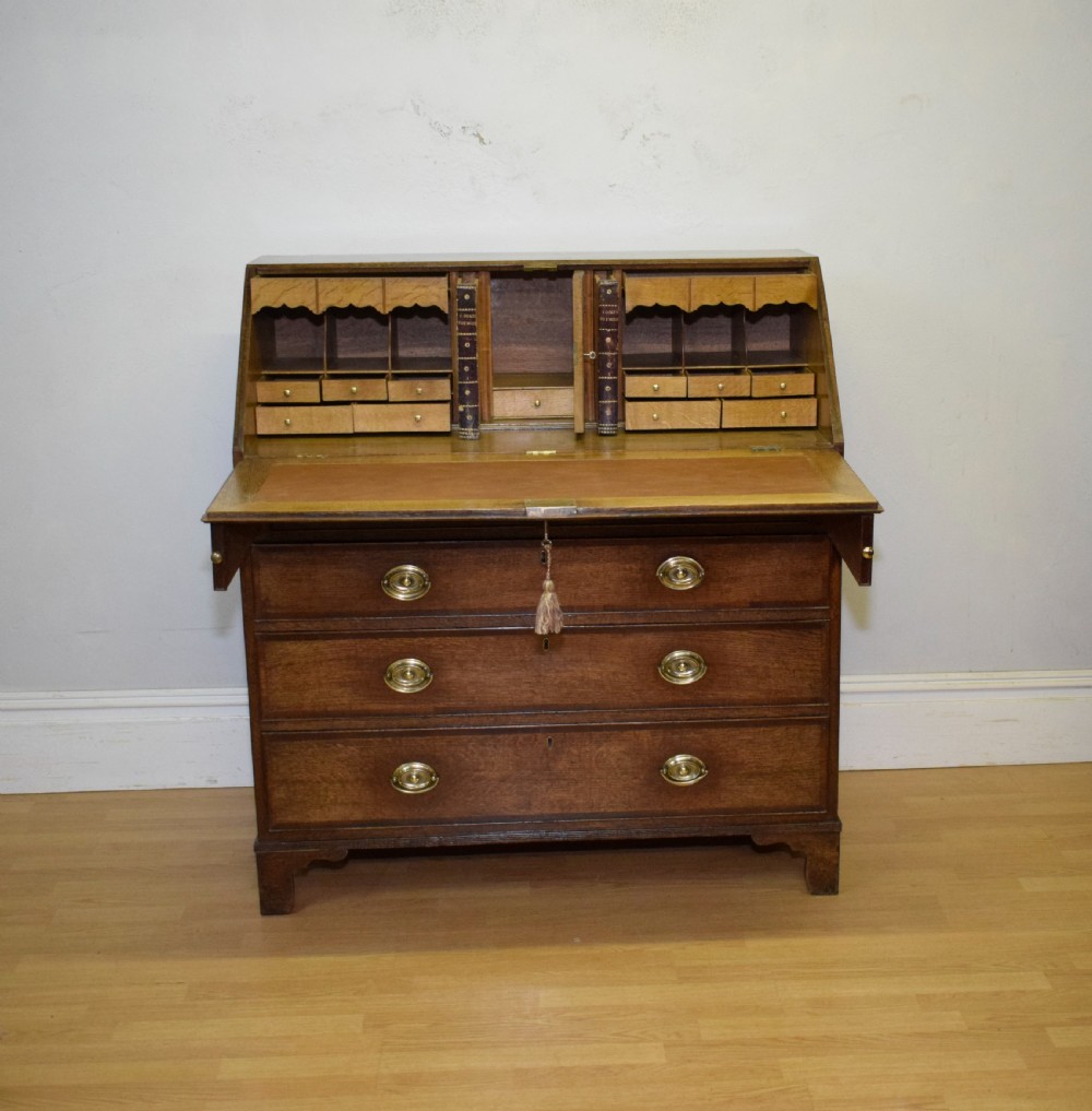 A Georgian oak bureau featuring a slanted fall-front lid above four graduated drawers fitted with brass oval handles and raised on bracket feet. View X