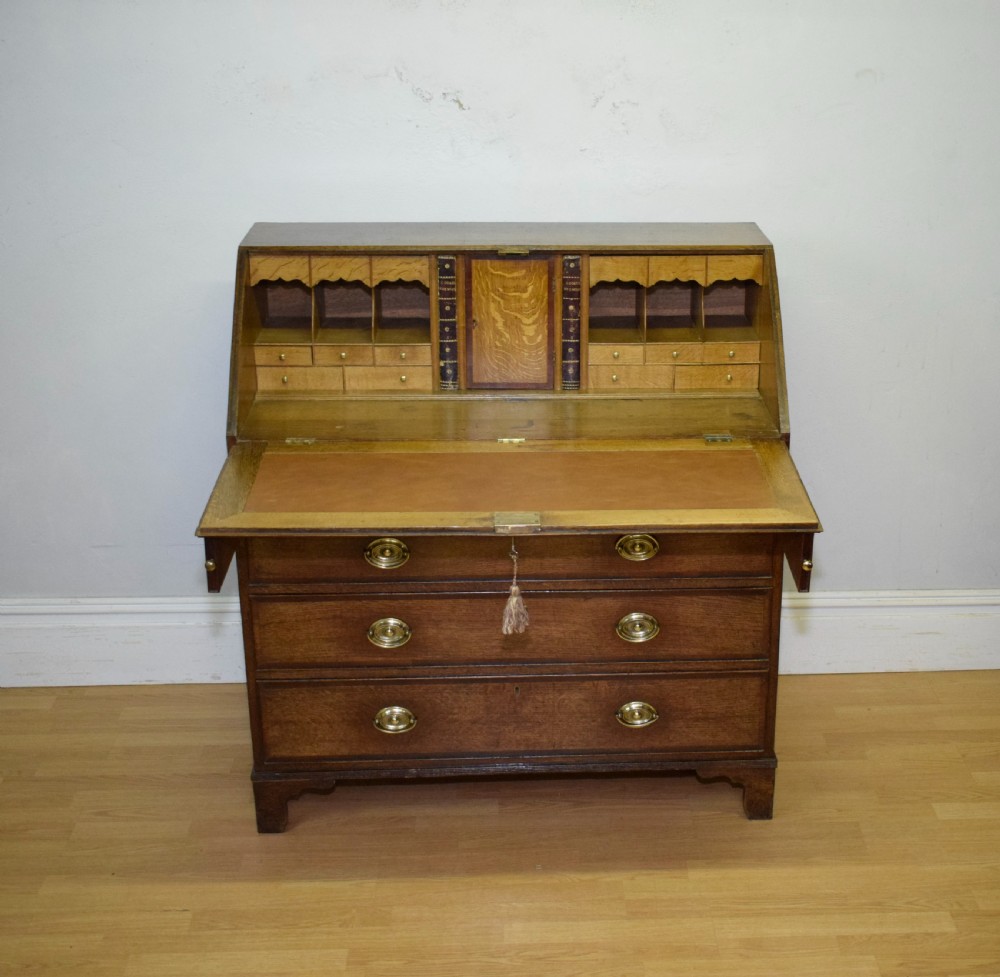 A Georgian oak bureau featuring a slanted fall-front lid above four graduated drawers fitted with brass oval handles and raised on bracket feet. View XII