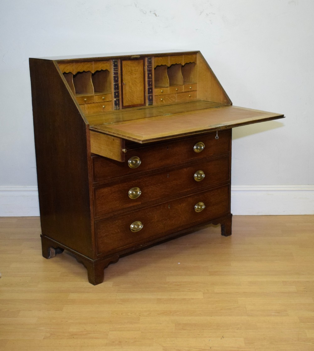 A Georgian oak bureau featuring a slanted fall-front lid above four graduated drawers fitted with brass oval handles and raised on bracket feet. View XV