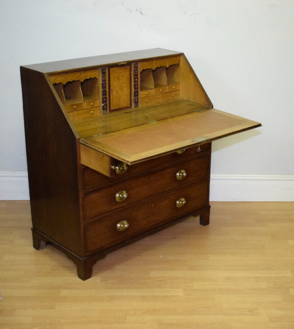 A Georgian oak bureau featuring a slanted fall-front lid above four graduated drawers fitted with brass oval handles and raised on bracket feet. View XVI