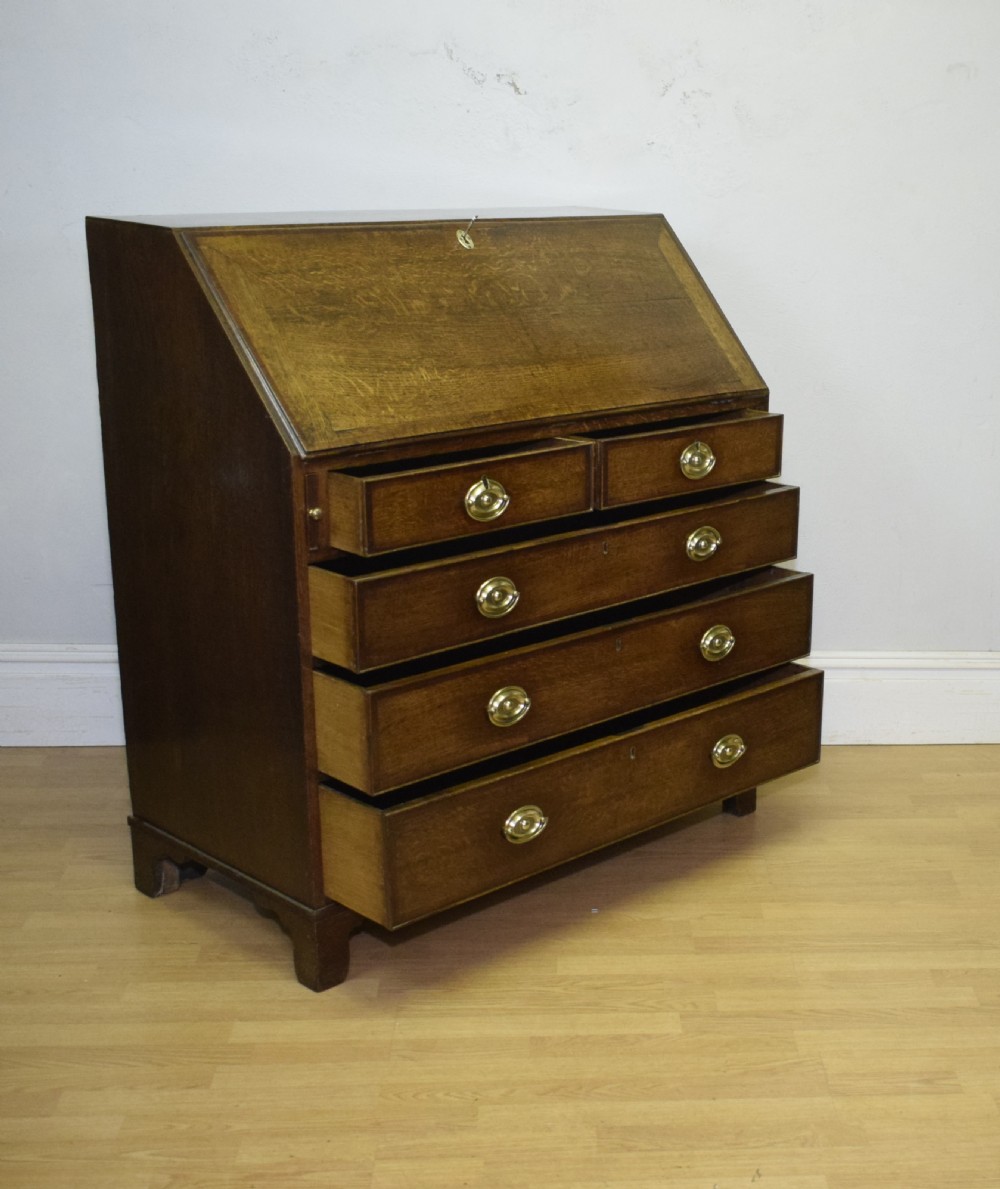 A Georgian oak bureau featuring a slanted fall-front lid above four graduated drawers fitted with brass oval handles and raised on bracket feet. View XVIII