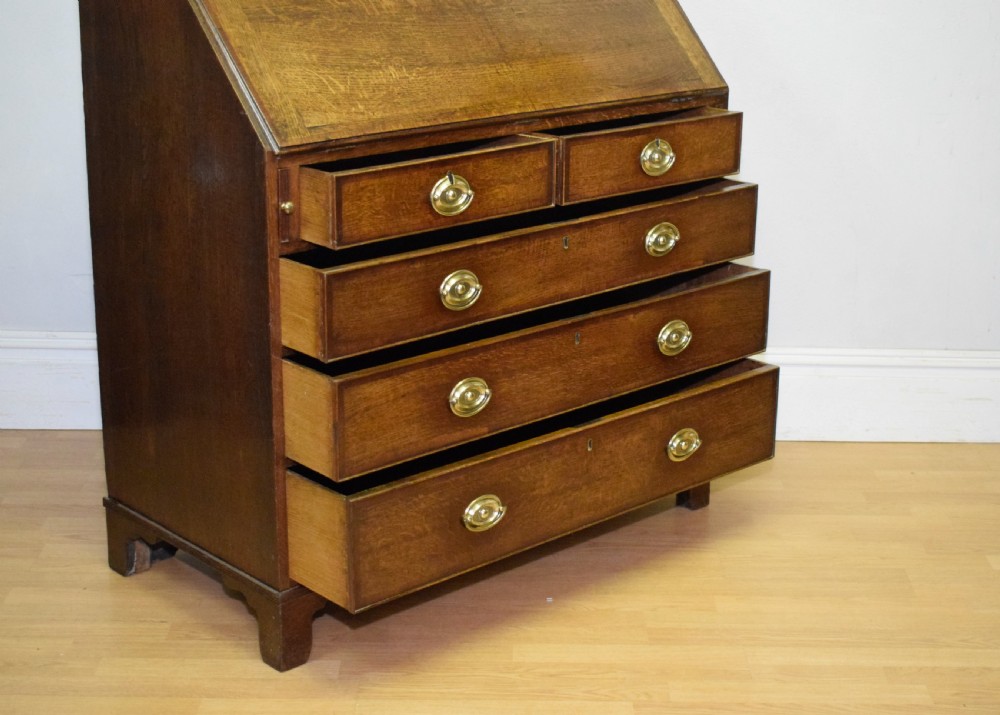A Georgian oak bureau featuring a slanted fall-front lid above four graduated drawers fitted with brass oval handles and raised on bracket feet. View XIX