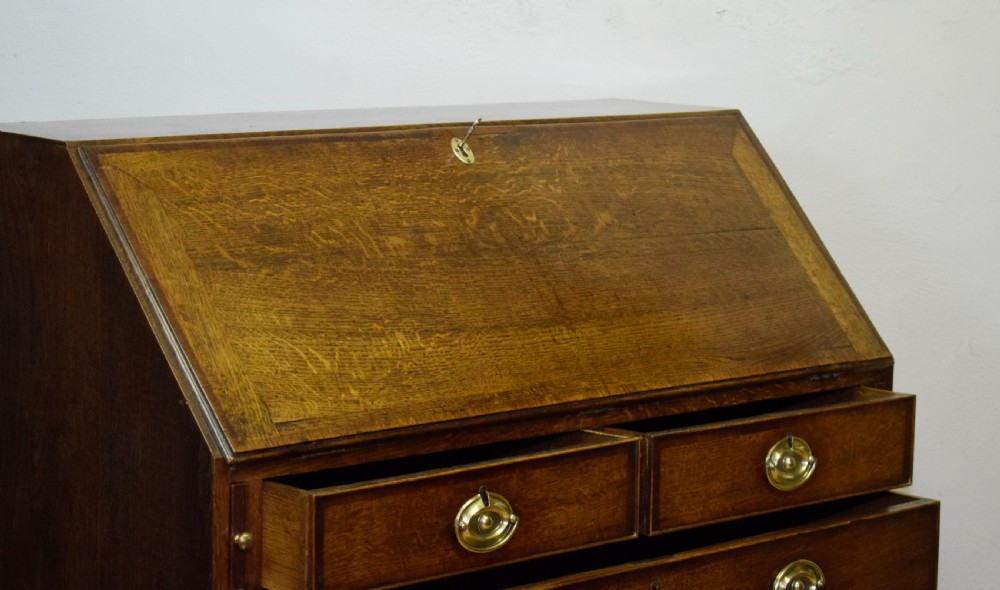 A Georgian oak bureau featuring a slanted fall-front lid above four graduated drawers fitted with brass oval handles and raised on bracket feet. View XX