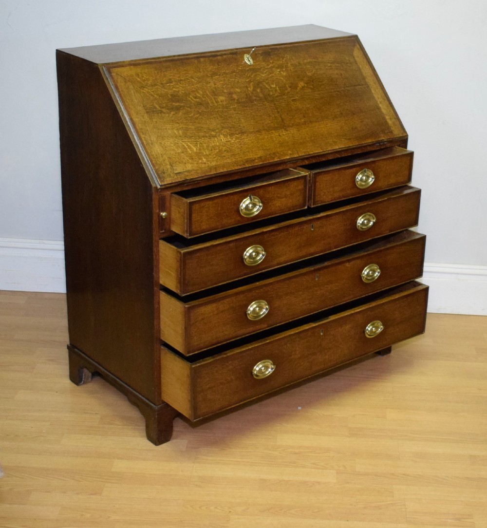 A Georgian oak bureau featuring a slanted fall-front lid above four graduated drawers fitted with brass oval handles and raised on bracket feet. View XXI