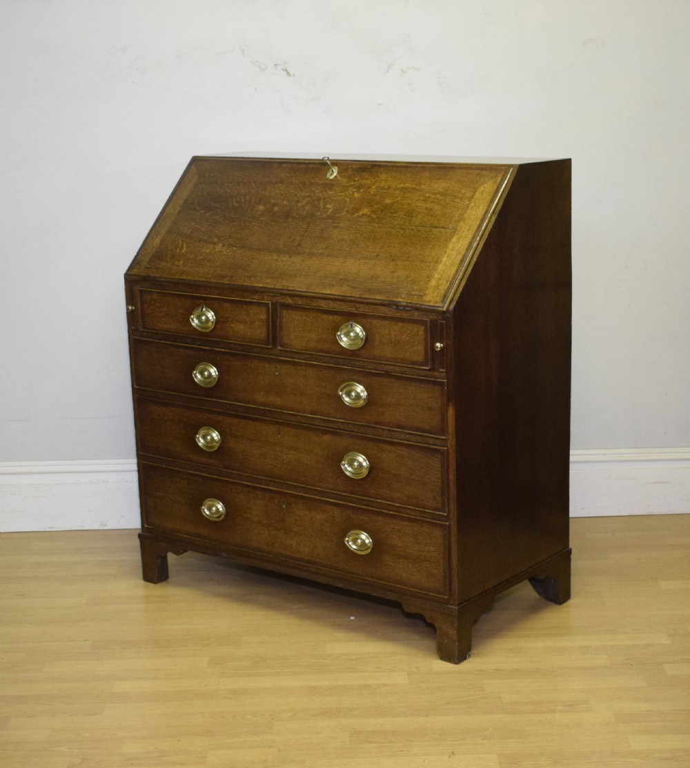 A Georgian oak bureau featuring a slanted fall-front lid above four graduated drawers fitted with brass oval handles and raised on bracket feet. View XXII