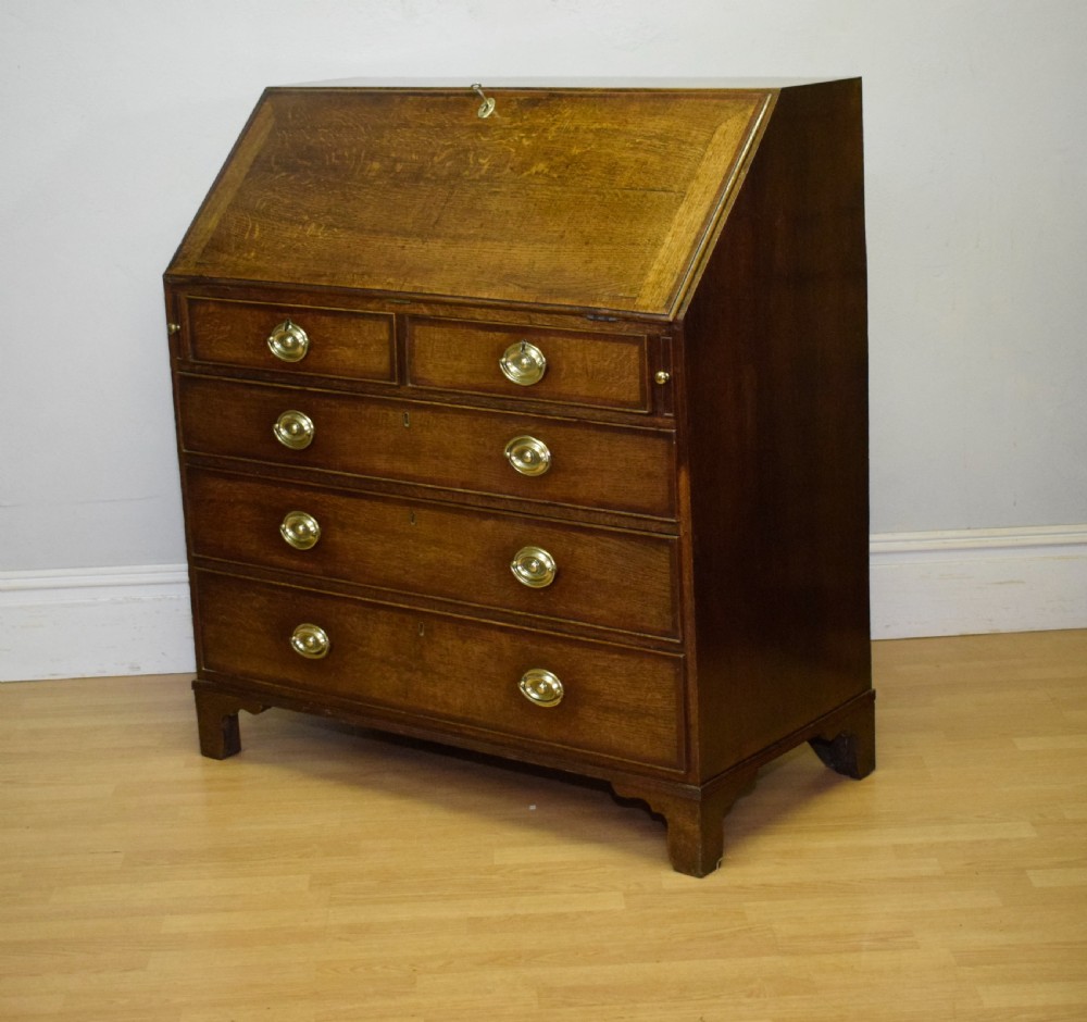 A Georgian oak bureau featuring a slanted fall-front lid above four graduated drawers fitted with brass oval handles and raised on bracket feet. View XXIII