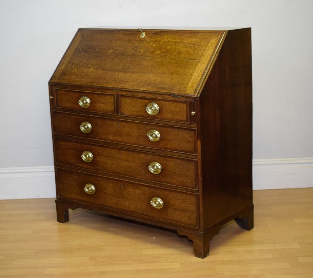 A Georgian oak bureau featuring a slanted fall-front lid above four graduated drawers fitted with brass oval handles and raised on bracket feet. View XXIV