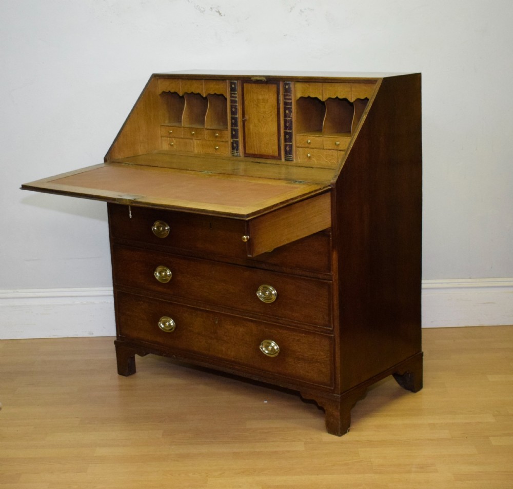 A Georgian oak bureau featuring a slanted fall-front lid above four graduated drawers fitted with brass oval handles and raised on bracket feet. View XXV